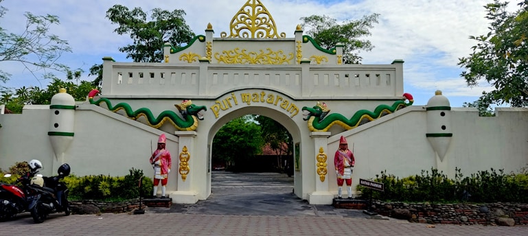 An ornate entrance gate features traditional architectural elements with decorative carvings and vibrant colors. Two statues of guards in traditional attire flank the archway. The gate is adorned with two dragon-like figures, and the name 'Puri Mataram' is inscribed in gold lettering. The setting includes trees and motorcycles parked nearby, adding a touch of everyday life to the scene.