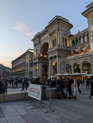 A bustling urban plaza with a large, ornate building featuring arches and columns. The façade is illuminated, hinting at a historical or significant site. People are gathered around, some walking, while others are stationary, possibly tourists or locals engaging in evening activities.