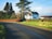 A picturesque countryside scene featuring a house surrounded by trees and bushes. A large tree stands prominently beside the house, and a curved road leads away, bordered by lush green grass. The sky is clear with a few clouds, lending a serene atmosphere.