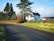 A picturesque countryside scene featuring a house surrounded by trees and bushes. A large tree stands prominently beside the house, and a curved road leads away, bordered by lush green grass. The sky is clear with a few clouds, lending a serene atmosphere.