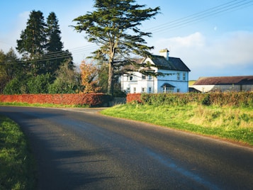 A picturesque countryside scene featuring a house surrounded by trees and bushes. A large tree stands prominently beside the house, and a curved road leads away, bordered by lush green grass. The sky is clear with a few clouds, lending a serene atmosphere.