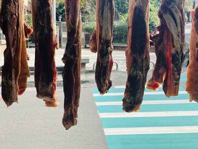 Several pieces of meat are hanging vertically in a row, suspended above a sidewalk with a striped crosswalk in the background. They appear to be drying or curing in the open air, and there is a mix of sunlight and shadow on the scene.