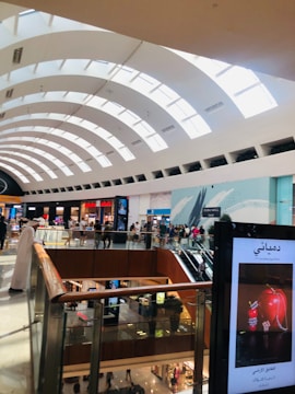 A spacious shopping mall with a high, curved ceiling featuring large skylights, allowing natural light to fill the area. The mall is two stories, with glass railings and various stores visible, including clothing and electronic shops. Shoppers are walking around, and there is a display screen featuring an advertisement.