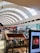 A spacious shopping mall with a high, curved ceiling featuring large skylights, allowing natural light to fill the area. The mall is two stories, with glass railings and various stores visible, including clothing and electronic shops. Shoppers are walking around, and there is a display screen featuring an advertisement.