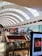 A spacious shopping mall with a high, curved ceiling featuring large skylights, allowing natural light to fill the area. The mall is two stories, with glass railings and various stores visible, including clothing and electronic shops. Shoppers are walking around, and there is a display screen featuring an advertisement.