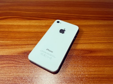 A white smartphone with the Apple logo on its back is placed at an angle on a wooden surface. The phone has a sleek design and reflective surface, emphasizing its minimalist aesthetic.