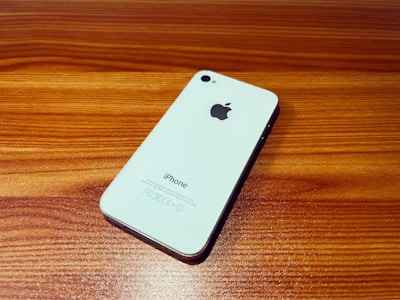 A white smartphone with the Apple logo on its back is placed at an angle on a wooden surface. The phone has a sleek design and reflective surface, emphasizing its minimalist aesthetic.