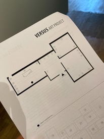 A hand holds a printed map or layout sheet labeled 'VERSUS ART PROJECT.' The paper features a grid-like floor plan designating areas with numbers and a depiction of a bathtub, likely part of an exhibition. The bottom section includes a legend with squares numbered from 18 to 29, and a black dot indicating a 'Wall Installation.'