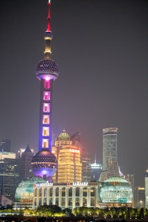 A nighttime cityscape showcasing a brightly illuminated futuristic tower with spherical segments, surrounded by other high-rise buildings. The architecture features a mix of modern and traditional elements with vibrant lighting.