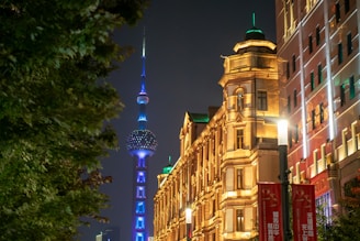 A nighttime cityscape featuring a brightly illuminated architectural tower, likely the Oriental Pearl Tower, glowing with blue and purple lights. Nearby, an ornately designed building with warm, yellow lighting contrasts with the modern tower. The street is lined with trees and additional buildings, with street lights and red banners visible.