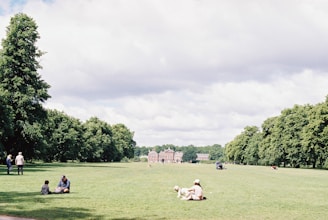 A large, open grassy park with scattered people enjoying the outdoors. Some people are sitting with a dog, while others are walking along tree-lined paths. A historic building is visible in the background surrounded by lush greenery and the sky is partly cloudy.