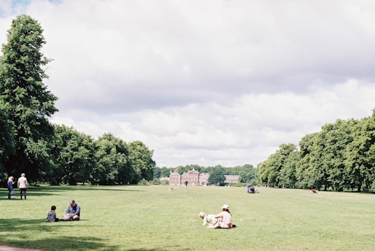 A large, open grassy park with scattered people enjoying the outdoors. Some people are sitting with a dog, while others are walking along tree-lined paths. A historic building is visible in the background surrounded by lush greenery and the sky is partly cloudy.