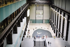 A large, modern indoor space with high ceilings and steel beams, resembling an art gallery or museum. The floor is a polished grey concrete, and a central black art installation is surrounded by people observing and interacting. Bright natural light enters through upper windows, creating an airy atmosphere.