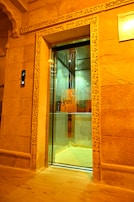 An ornate elevator door set in a decorative stone wall, with intricate carvings framing the entrance. The elevator interior features a mirrored back wall and a reflective floor. A floor indicator on the left shows the number two.