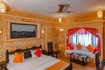 The image depicts a well-decorated, cozy bedroom that features a double bed with white bedding, accented by orange and red pillows. To the right is a window with light blue curtains framed by red drapes, and a seating area below it with matching cushions. The room has beige stone walls with an intricate design, and ornate lighting fixtures hanging from the ceiling and mounted on the walls. A large black and white picture hangs above the bed, and there is a ceiling fan in the center.