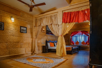 A richly decorated room with ornate carved stone walls, featuring a large bed adorned with red and orange throw pillows. The room includes a ceiling fan, framed pictures on the wall, and curtains that elegantly drape around the bed area. A patterned carpet lies on the polished floor, and ambient lighting creates a warm atmosphere.