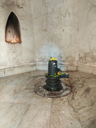 A marble floor surrounds a dark stone structure adorned with flowers and green leaves, typically indicative of a religious or spiritual setting. The walls feature graffiti, with a niche housing a small flame or candle.