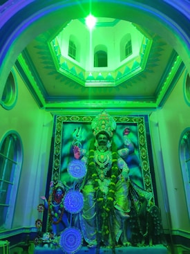 A vibrant, illuminated shrine featuring a large, ornate statue of a deity with multiple arms. The setting is adorned with intricate decorations and floral garlands. Green lighting casts a mystical glow over the entire scene, accentuating the architectural details of the surrounding space.