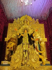 A detailed and ornate idol of a goddess is richly decorated with gold and intricate patterns. The deity is adorned with an ornate crown and jewelry and is placed in front of an elaborately designed backdrop featuring detailed carvings and embellishments. A lion, associated with the goddess, is depicted beside her with a fierce expression. Above, a chandelier with glass or crystal elements adds grandeur to the scene, while rich maroon curtains frame the setting.