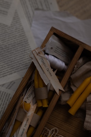 A wooden box filled with hand-rolled yellow candles secured with paper bands and handwritten labels. The box is placed amidst open books or paper sheets with text. There are additional candles and string visible, giving a warm and rustic atmosphere.