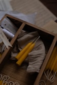 A wooden box divided into sections contains several small hand-rolled yellow beeswax candles, some resting on a piece of natural-colored fabric. Papers and possibly tags are scattered nearby.