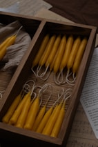 A wooden box containing neatly arranged yellow taper candles, surrounded by sheets of paper with music and text. The candles are aligned in two sections, and some are resting on a neutral-colored fabric inside the box.