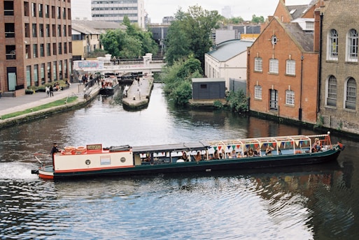 A canal boat filled with people is traveling through a narrow waterway bordered by buildings and greenery. On the left, modern brick buildings rise along the canal, while pedestrians are gathered near the canal edge. On the right, older brick buildings with arched windows add a historic feel. In the background, a bridge crosses the canal and is adorned with graffiti.