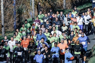 A large group of runners participating in a marathon or organized run along a tree-lined road. The participants wear diverse athletic attire in various colors, and many have race numbers attached to their shirts. The surrounding environment includes a grassy area and trees with autumn foliage.