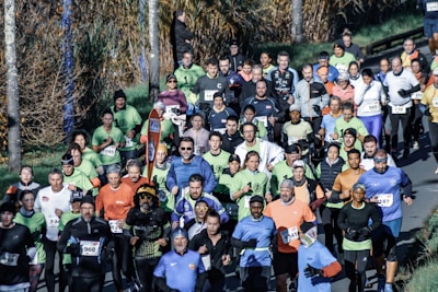 A large group of runners participating in a marathon or organized run along a tree-lined road. The participants wear diverse athletic attire in various colors, and many have race numbers attached to their shirts. The surrounding environment includes a grassy area and trees with autumn foliage.