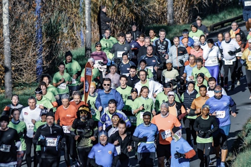 A large group of runners participating in a marathon or organized run along a tree-lined road. The participants wear diverse athletic attire in various colors, and many have race numbers attached to their shirts. The surrounding environment includes a grassy area and trees with autumn foliage.