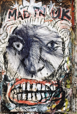 A vivid and chaotic graffiti-style artwork features a large, distorted face with exaggerated eyes and a mouth full of teeth. The words 'MAD IN UK' are prominently painted above the face, surrounded by abstract lines and splatters.