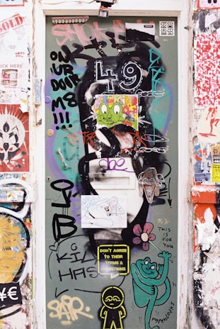 A door covered in vibrant graffiti and stickers, including various artistic designs and messages such as 'Don't agree to their terms & conditions' and 'This is for you.' The numbers '4 9' are prominently displayed, along with cartoon-like characters and colorful illustrations. Surrounding the door are more layers of urban street art and posters.