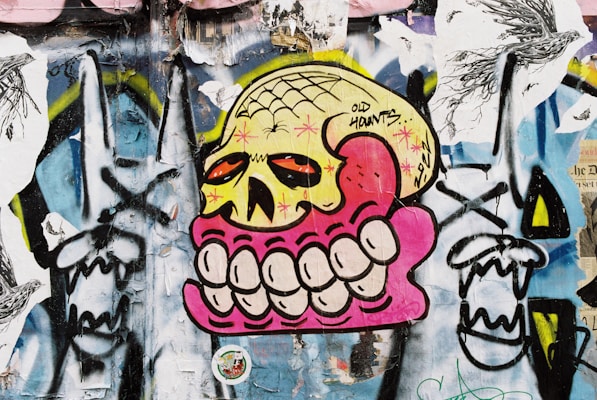 A vibrant and eclectic piece of street art featuring a large stylized skull with exaggerated teeth, rendered in bold colors against a textured wall. The skull is yellow with red eyes, surrounded by a pink outline, and the teeth are oversized and prominent. The background is a collage of various graffiti elements, including swirling black and white patterns and abstract shapes, creating a dynamic and chaotic visual effect.