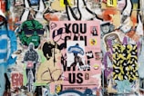 A collage of vibrant street art and posters covers a wall. Multiple layers of graffiti and decals include depictions of stylized faces, a green figure wearing sunglasses, a person on a bicycle, and a large pink poster with bold text. Various stickers and smaller images are scattered throughout, contributing to an eclectic and chaotic aesthetic.