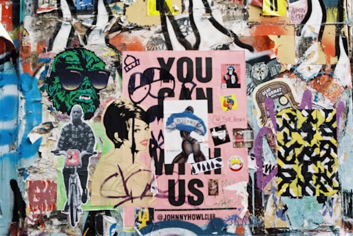 A collage of vibrant street art and posters covers a wall. Multiple layers of graffiti and decals include depictions of stylized faces, a green figure wearing sunglasses, a person on a bicycle, and a large pink poster with bold text. Various stickers and smaller images are scattered throughout, contributing to an eclectic and chaotic aesthetic.