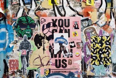 A collage of vibrant street art and posters covers a wall. Multiple layers of graffiti and decals include depictions of stylized faces, a green figure wearing sunglasses, a person on a bicycle, and a large pink poster with bold text. Various stickers and smaller images are scattered throughout, contributing to an eclectic and chaotic aesthetic.