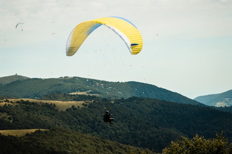 A person is paragliding over lush green hills and forests under a partly cloudy sky. The large, yellow and white paraglider contrasts against the natural landscape, creating a sense of adventure and freedom. Other paragliders are visible in the distance, adding to the dynamic scene.