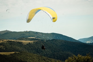 A person is paragliding over lush green hills and forests under a partly cloudy sky. The large, yellow and white paraglider contrasts against the natural landscape, creating a sense of adventure and freedom. Other paragliders are visible in the distance, adding to the dynamic scene.