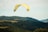 A person is paragliding over lush green hills and forests under a partly cloudy sky. The large, yellow and white paraglider contrasts against the natural landscape, creating a sense of adventure and freedom. Other paragliders are visible in the distance, adding to the dynamic scene.