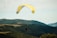 A person is paragliding over lush green hills and forests under a partly cloudy sky. The large, yellow and white paraglider contrasts against the natural landscape, creating a sense of adventure and freedom. Other paragliders are visible in the distance, adding to the dynamic scene.