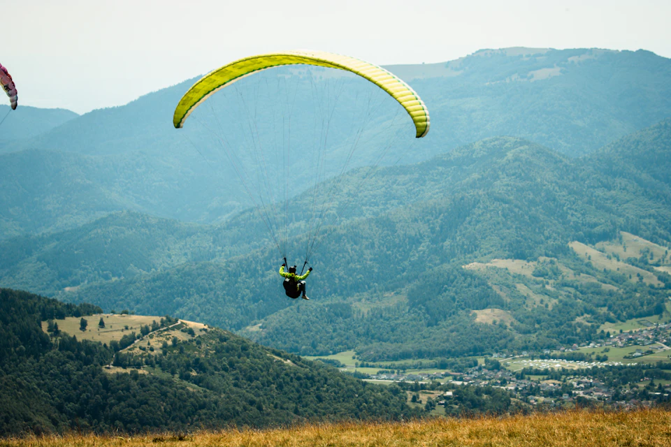 The image depicts a person paragliding over a verdant valley, with their body suspended in mid-air and attached to a vibrant yellow parachute. The paraglider is positioned centrally in the frame, creating a sense of balance between the soaring individual and the expansive landscape below. In the distance, mountains rise against an azure sky, adding depth and grandeur to the scene.