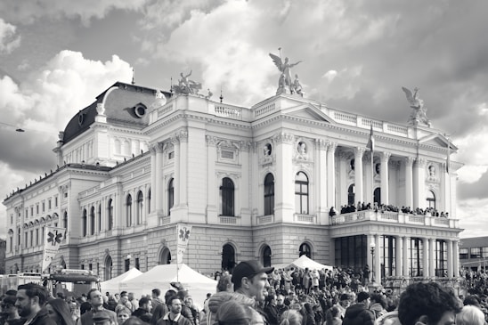 A large crowd is gathered in front of a grand classical building which features ornate architectural details such as columns and statues. The building is likely of historical or cultural significance. People appear engaged and possibly participating in an event or protest. The image is in black and white.