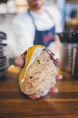 A person holds a pita bread filled with grilled meat and topped with white and yellow sauces. The background is slightly blurred, suggesting a kitchen or restaurant setting. The hand is prominently featured, holding the dish over a wooden surface.