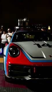 A sporty red and white car with blue racing stripes is prominently displayed in a nighttime setting. The car's front design is sleek with a visible number '14' on its hood. In the background, a group of people, some wearing hats, are gathered, observing the car. The scene is dimly lit, creating a focus on the car's vivid colors.