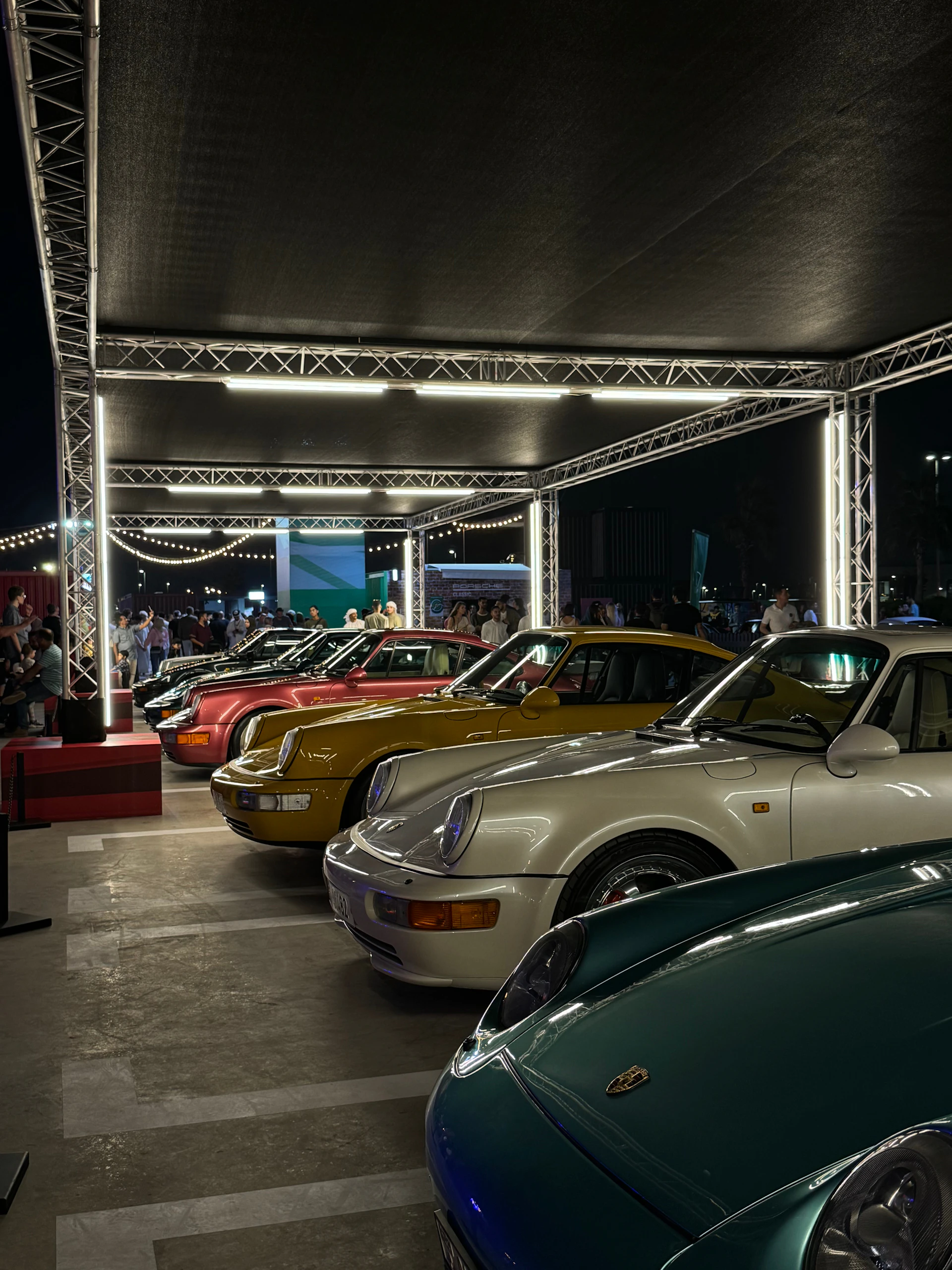 Evening scene of classic cars parked under string lights with people enjoying the warm atmosphere of the Vindimas gathering.