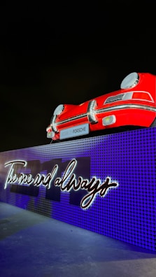 A large decorative display featuring the front of a classic red Porsche prominently mounted atop a patterned blue and black structure. The sign below it reads 'Icons of Porsche' and features the luminous words 'Then and always' in a stylish script font.