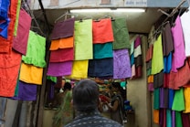 An assortment of brightly colored fabrics hangs on display in front of a small shop. The fabrics are neatly arranged in rows, showcasing a variety of vivid colors such as red, green, purple, and orange. People inside the shop are browsing and examining the fabrics, while a person in the foreground is seen from behind observing the display.