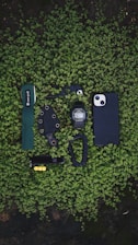 A collection of everyday carry items neatly arranged on a bed of small green leaves. The items include a green wrist strap with 'snow peak' branding, a black multi-tool, a black digital wristwatch, a black smartphone case, a knife with a black sheath, a black carabiner, and a black Swiss army knife with yellow accents.