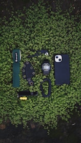 A collection of everyday carry items neatly arranged on a bed of small green leaves. The items include a green wrist strap with 'snow peak' branding, a black multi-tool, a black digital wristwatch, a black smartphone case, a knife with a black sheath, a black carabiner, and a black Swiss army knife with yellow accents.