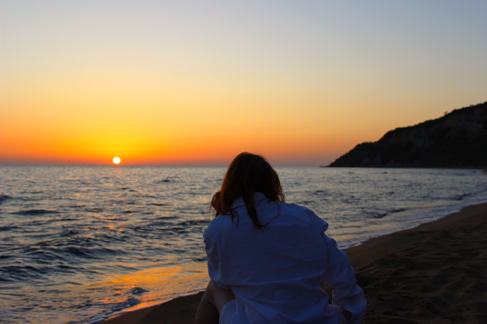 uma mulher sentada em uma praia observando o pôr do sol